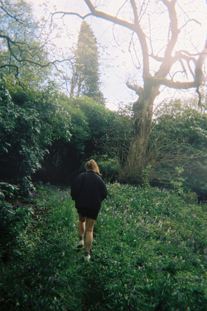 A person in a dark jacket walking through a green, overgrown garden toward a large bare tree