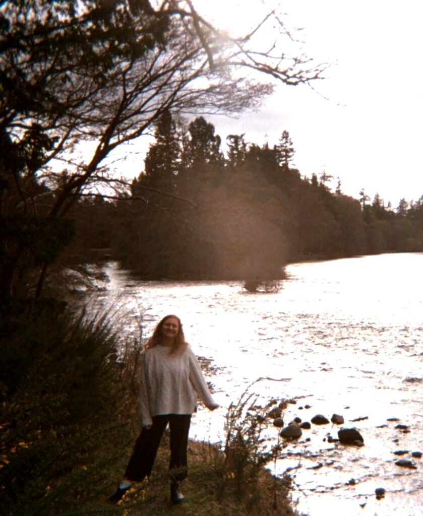 Kailee standing on a rocky riverbank with trees along the far shore