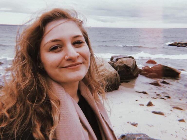 Kailee smiling on a windswept Cape Cod beach, with waves and rocks behind her
