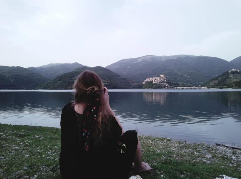 Kailee from behind, sitting on the grass by a calm lake in Italy, looking out at green mountains and a hilltop village