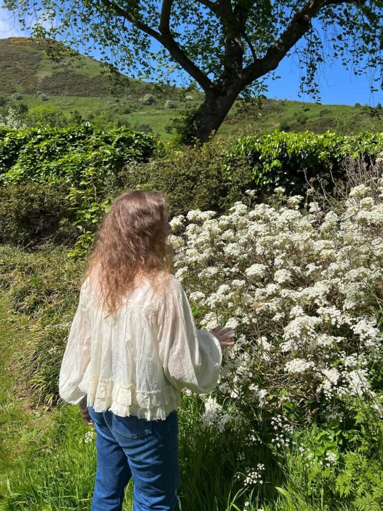 Kailee from behind, in a white blouse, touching a bush of white blossom beneath a tree and a green hill in Scotland