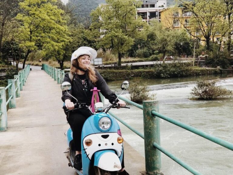 Kailee riding a pale blue scooter across a footbridge over a river in China, smiling in a white helmet
